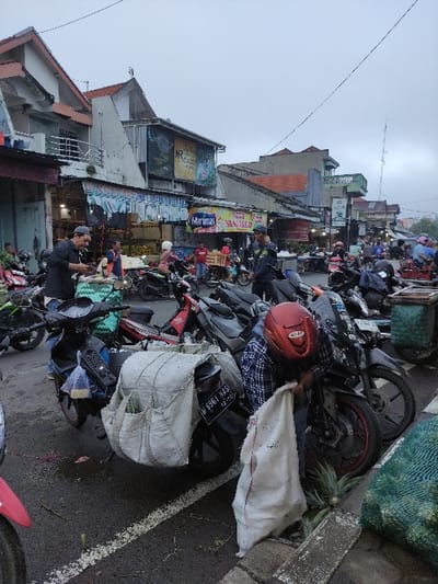 A man in a plaid shirt and red helmet is loading a large white sack onto a motorcycle in Mojokerto, Indonesia. The motorcycle is parked in front of several other motorcycles, all of which are parked in front of a row of buildings. There are several other people in the scene, but they are not in focus. The buildings are all in a row, and they are all painted in a variety of colors. The road is paved and there are white lines painted on it. The sky is cloudy and overcast. It is daytime.