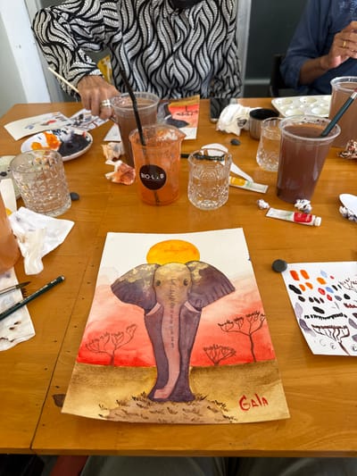 A top-down medium shot captures a wooden table filled with art supplies, suggesting an art class or workshop. In the foreground, a watercolor painting of an elephant against a sunset landscape is laid out. The elephant is depicted in shades of gray and purple, with golden accents on its ears and forehead. The background features a vibrant orange sun and silhouette trees in shades of red and brown. The signature "Gaia" in red is visible in the bottom right corner of the painting.
Scattered around the painting are various art materials: several clear plastic cups containing brown and orange liquids, one with a "BIO-LAB COFFEE & OFFICE" label; a small white palette with dabs of orange and black paint; a glass of water; a tube of red paint; crumpled paper towels; and a green drawing pencil. In the background, two individuals are partially visible. One person on the left wears a black and white patterned shirt and holds a paintbrush, appearing to be painting. Another person on the right wears a dark blue shirt and has their hands near art supplies.
The overall impression is of a creative and engaging activity taking place in a well-lit indoor space with a wooden table as the central element. The setting is not definitively Siem Reap, Cambodia, as there are no specific visual cues to indicate this location.