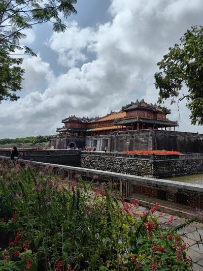 This image captures the Ngọ Môn Gate (Meridian Gate) of the Imperial City of Huế, located in Phú Xuân Ward, Vietnam, under an overcast sky with diffused light.

In the foreground, a dense bed of green plants with scattered red and purple flowers occupies the lower left. A light-colored concrete pathway with decorative balustrades runs horizontally across the mid-foreground, bordering a dark body of water, likely a moat. Beyond the moat, a robust, dark grey stone wall, constructed from stacked rough stones, forms a bank, featuring a rectangular planter box filled with vibrant red flowers in the mid-right.

The central architectural subject is the multi-tiered Ngọ Môn Gate, presented as a prominent structure built on a raised dark grey stone platform. The gate features traditional Vietnamese design elements, including elaborate dark-tiled roofs with decorative finials, visible red support columns, and lighter orange-ttiled upper sections. A large arched entrance is centrally positioned within the stone platform, with additional smaller arched or rectangular openings visible. Approximately 15-20 individuals, appearing to be visitors, are congregated on the platform and near the gate entrance. A single individual in dark clothing stands on a pathway to the far left, facing the gate.

A stone bridge crosses the moat, providing access to the gate's platform. Green tree foliage partially frames the image from the upper left and right corners. A continuous band of green trees lines the top of the distant wall, which extends horizontally behind the main gate structure.