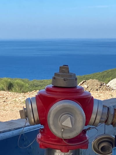 A red fire hydrant is positioned in the foreground, facing the viewer, with a large body of blue water and a hazy horizon in the background. The hydrant features silver accents around its various nozzles and a silver cap on top. A thin wire is attached to the hydrant's left nozzle, looping downwards. The hydrant sits on a concrete surface, which extends to the left and right. Behind the hydrant, a gently sloping hill is covered in sparse green vegetation and rocky terrain. The sky above is a clear, light blue, fading into a lighter shade towards the horizon. The scene appears to be outdoors, possibly in a coastal or hilly area. The text "CE 1404" is visible at the base of the hydrant.