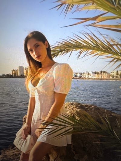 The media file depicts a young woman with long, dark hair leaning against a rocky surface near the water. She is wearing a light-colored, possibly white, dress with short sleeves and a gathered detail on the bodice. Her left arm is bent and resting on her lap, while her right arm is extended outwards. Her expression appears relaxed and contemplative.
The scene is set outdoors, with a body of water, likely the sea or a large lake, occupying the background. The water has a rippled texture, suggesting a gentle breeze. In the distance, across the water, a line of buildings with a light color palette is visible, hinting at a coastal town or resort. Palm fronds, casting shadows, frame the top and right sides of the image, indicating a warm climate. The bright sky suggests it is daytime, likely sunny. The overall mood is peaceful and serene, characteristic of a vacation or leisure setting. There is no visible text in the image.