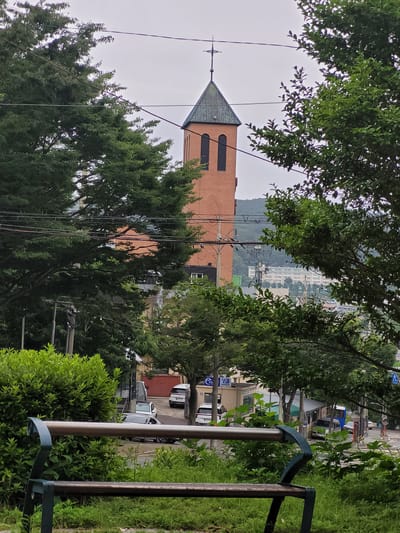 An outdoor, eye-level, medium shot captures a church tower with a green, sloped roof and a cross on top, made of red brick, set amidst lush green trees. The tower has two tall, arched, dark windows. Power lines crisscross the frame, and a hazy, distant view of buildings and hills suggests an urban or suburban setting, possibly in Busan, South Korea. In the foreground, a dark green metal park bench with a wooden seat sits on a grassy incline. A street with several parked cars, including a silver sedan and white vans, is visible beyond the trees, along with a building that appears to be a small business or service booth. The sky is overcast.