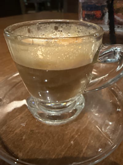 The image shows a glass mug with a handle, partially filled with a beverage, likely coffee or a similar foamy drink. The mug sits on a clear glass saucer placed on a wooden surface. The environment appears to be indoors. Though I do not have enough information to determine a specific location in Dania Beach.
