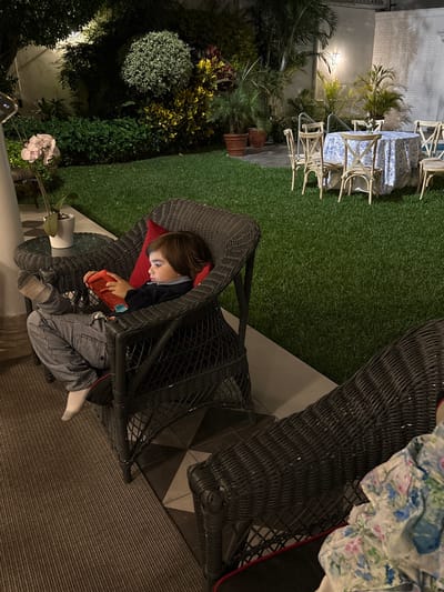 A child is sitting in a black wicker chair, holding a red electronic device, possibly a gaming console. The child wears striped pants and socks. A red pillow supports the child's back. The chair is on a tiled surface next to a textured rug. The scene is set outdoors, with lush green grass and various plants. A table and chairs with a patterned tablecloth are visible in the background. A potted plant with white flowers sits near the chair. The location is likely a residential patio or garden in San Isidro, Peru.