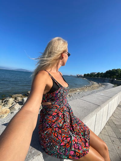A blonde woman with her hair blowing in the wind, wearing sunglasses and a colorful dress, is sitting on a stone wall overlooking a body of water. The scene appears to be a waterfront promenade in Sementsovka, Russia, with a rocky shore and buildings visible in the distance. The sky is clear and blue, indicating a sunny day. The woman is looking out at the water, with her arm extended towards the camera as if taking a selfie. There are a few other people walking on the beach in the background.