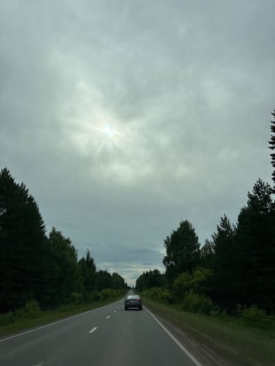 A two-lane asphalt road stretches into the distance, flanked by dense coniferous and deciduous trees. The sky is overcast with the sun partially obscured by clouds, creating a bright halo effect. A dark-colored sedan is traveling away from the viewer in the right lane. The road surface appears to be in good condition with white dashed lines marking the lanes. The environment suggests a rural or semi-rural setting, likely within the city of Забегалово, Russia, given the context.