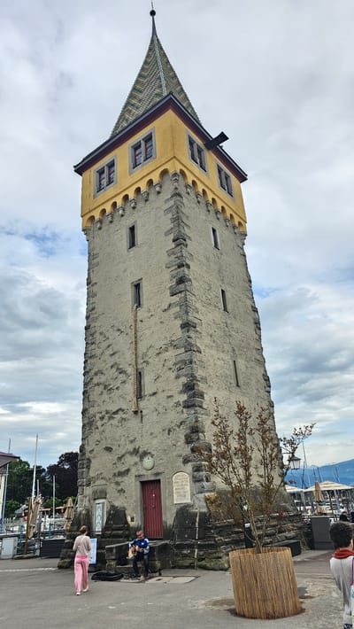 The image depicts the Mangturm, a medieval tower in Lindau (Bodensee), Germany. The tower is constructed of light-colored stone and has a distinctive pointed roof covered in colorful tiles. The upper portion of the tower's facade is painted yellow and features a series of windows. The lower portion of the tower has a wooden door. A tree is planted in a rectangular pot in front of the tower. Two people are on the ground nearby, seemingly interacting with each other. The background shows a view of the Lindau harbor with boats and a cloudy sky.