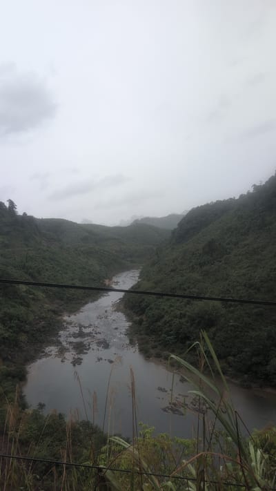 The image depicts a natural landscape in Trường Sơn Commune, Vietnam, on an overcast day. A wide, calm river flows through a valley flanked by steep, densely vegetated hills. The riverbed is visible in places, with scattered rocks and shallow areas. In the foreground, tall grasses and reeds grow along the riverbank, partially obscuring the view. Two horizontal cables, possibly from a power line or bridge, stretch across the middle ground of the image. The distant hills are shrouded in a light mist, creating a sense of depth and atmospheric haze. There are no people or animals visible in the scene. The overall impression is one of a remote and undeveloped natural environment.