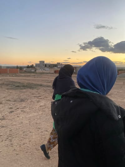 Two individuals are captured in a slightly tilted outdoor shot. The person closest to the camera wears a black jacket, patterned pants, and black shoes, with a glimpse of a green shirt underneath. The other individual is dressed in a blue head covering. The setting includes a sky with a mix of blue and golden hues, possibly during sunrise or sunset, and a landscape featuring sparse buildings and a dirt ground. The shot is somewhat blurred.