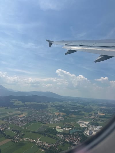 The image captures an aerial view from an airplane window, looking down at a landscape that includes the village of Siezenheim, Austria. The prominent subject is the wing of the airplane, which occupies the left side of the frame. It is light gray and angled, with visible flaps and supports. The rest of the scene is dominated by the sky and the ground below.
The sky is a bright blue with scattered white clouds, some of which are large and puffy, suggesting a fair weather day. The sun appears to be high in the sky, indicating it is likely daytime, possibly late morning or early afternoon.
Below the airplane wing, the landscape is a patchwork of green fields, some with geometric patterns that suggest agricultural use, possibly including vineyards or orchards. A village with red-tiled roofs is visible, nestled among the fields and trees. A body of water, possibly a river or a lake, is also present. In the distance, mountains rise, their peaks shrouded in a hazy blue, suggesting they are further away or the atmosphere is slightly misty. There are also some man-made structures, including what appears to be a commercial building or industrial complex with a large parking lot, and a residential area with houses.
The image is oriented vertically, suggesting it was taken by holding the camera in a portrait orientation. There is no visible text in the image. The overall impression is one of travel and a scenic view from above.
