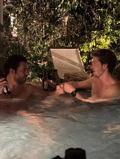 At night in Valle de Bravo, Mexico, two adult males are partially submerged in a bubbling outdoor hot tub. The male on the left, with dark hair and a beard, holds a dark-colored bottle in his right hand. He is engaged in a fist bump with the male on the right, who has lighter hair and wears a dark smartwatch on his left wrist. A second dark-colored bottle is visible resting near the left male's arm. In the background, dense green foliage is illuminated by artificial light, behind which an outdoor lounge chair with a folded item of fabric is present.