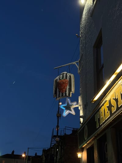 A rotated, nighttime image features a dark blue sky. In the foreground, a striped object with a large strawberry on it is suspended. Next to it is a glowing blue star. Above, the underside of a building is visible, illuminated by a light fixture. A sign reading "The Fest" is present, along with a set of stairs and additional lighting fixtures on the building.