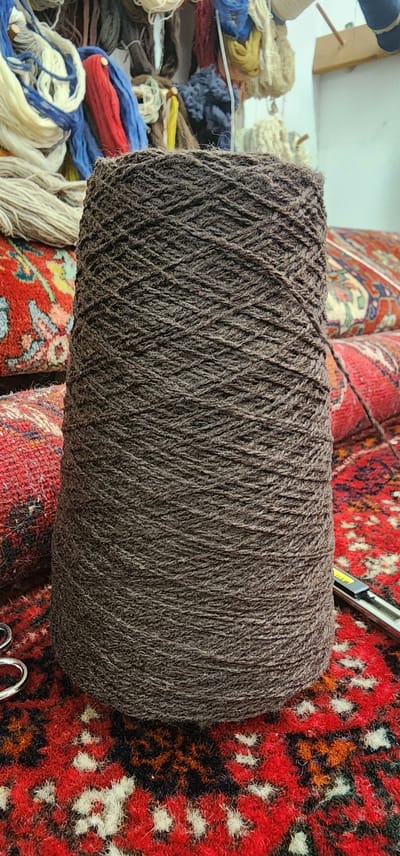 The image shows a close-up view of a large spool of dark brown yarn, centrally placed on a red rug with an intricate pattern. The yarn is tightly wound around a conical core, revealing the textured strands that make up its composition. To the left of the spool, stacks of colorful yarn in shades of white, blue, red, and yellow are visible, along with what appear to be portions of other rugs or textile materials. A small, silver object, possibly scissors or a craft knife, rests on the rug to the right of the spool. The background is slightly out of focus, but suggests an indoor setting with more yarn and textile elements, and a wooden beam with spools of yarn is visible in the upper right corner. The scene is devoid of any people. There is no information within the image to indicate a specific location in the city of Lod, Israel.