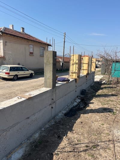 The image depicts an outdoor construction scene in Zlatovrah, Bulgaria, under a clear blue sky. In the foreground, a concrete wall extends diagonally from the lower-left to the mid-right. Vertical steel reinforcing bars protrude from the top of the concrete wall at regular intervals. Several sections of wooden formwork, containing vertical rebar, are erected along the wall, prepared for pouring additional concrete columns. One finished concrete column stands upright on the left portion of the wall. A narrow trench, containing excavated dirt and dark plastic sheeting, runs along the base of the wall on the viewer's side. To the left of the concrete wall, a light-colored station wagon, bearing Bulgarian license plate "PB 1424 CT," is parked on a dirt surface. Behind the vehicle, a two-story, light-colored residential building with a red-tiled roof and two chimneys is visible; one window on this building is boarded. Overhead power lines span the upper midground, supported by a wooden utility pole. The background shows additional distant buildings and a clear horizon with faint hills. No individuals are present in the scene.