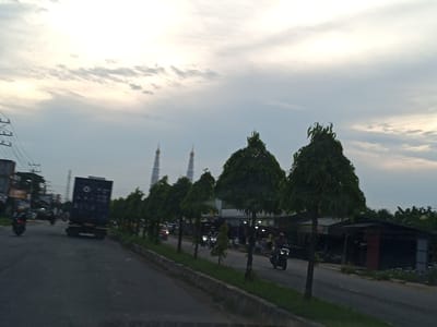 The image depicts a street scene in Payacut, Indonesia, at dusk. The sky is overcast with scattered clouds, suggesting a dimming light. On the left side of the road, a large, dark shipping container truck is visible, traveling forward. Several motorcycles are also on the road; one in the foreground on the left and others further down the street, with one rider clearly visible on the right. Lined along the right side of the road is a row of tall, conical trees. In the background, beyond the trees, the silhouettes of two mosque minarets are prominent against the sky. To the far right, a dark-colored building with an open front, possibly a shop or market stall, is present, with some structures visible behind it. The overall atmosphere suggests a typical evening on a busy street with ongoing traffic and the presence of religious structures.