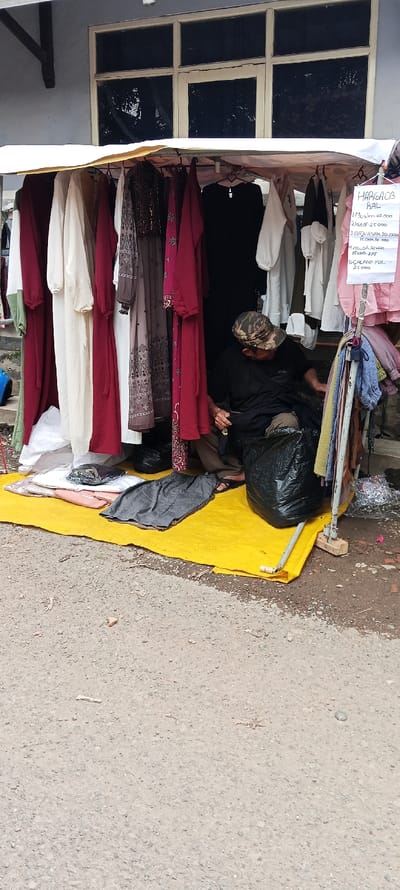 A man is sitting on a yellow mat in front of a stall with clothes hanging from a canopy. The stall is located in front of a building with a large window. The man is wearing a black shirt and black pants. He is looking at the ground. There is a black garbage bag next to him. The clothes hanging from the canopy are mostly long dresses and shirts, and there are some towels hanging on a rack behind the man. There is a sign hanging from the canopy that says "Harga 08" and lists prices for different types of clothing. The stall is located in front of a building in the city of Kutawaringin, Indonesia, at the coordinates -6.974 latitude and 107.519 longitude. The ground is covered in gravel and dirt. The building is painted a light blue color. The window in the building is made of glass and metal.