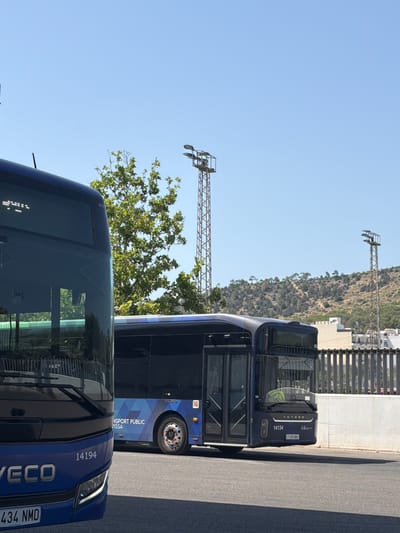 Two blue Iveco buses are parked side-by-side on a paved road, under a clear blue sky. The bus in the foreground, on the left, is a double-decker bus, and only its front half is visible. The bus behind it, on the right, is a single-decker bus. The front of the single-decker bus displays the number "14134" and the license plate "7498 NNV". The text "YUTONG" is also visible on the front of this bus, along with a logo. The side of the single-decker bus has text that reads "TRANSPORT PUBLIC", "VISSA", and the number "14134". The sun is bright, suggesting it is daytime, and the weather is clear. In the background, a steep, tree-covered hillside is visible, with some buildings at its base and top. There are also metal structures that appear to be lighting rigs or pylons. The setting appears to be a road or parking area, possibly near a transportation hub, in a Mediterranean climate. There are no people visible in the image.