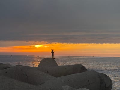 This is a photograph of a seascape at sunset in Jeju, South Korea. The foreground is dominated by large, grey concrete tetrapods, commonly used as breakwaters. Beyond the tetrapods, the ocean stretches to the horizon, reflecting the warm hues of the setting sun. Several small boats are visible on the water, some with lights illuminated, suggesting they are either fishing or traveling.

In the middle ground, a lighthouse stands on a breakwater. The lighthouse is cylindrical with a dark upper section and a yellow base, and it has a navigational light at its top. The sky above is a dramatic gradient of colors, transitioning from a deep grey at the top to vibrant oranges and yellows near the horizon, where the sun is setting. Thin clouds are scattered across the sky, catching the last rays of sunlight.

The overall mood of the image is serene and peaceful, capturing the beauty of a coastal sunset. The time of day is clearly sunset, indicated by the low angle of the sun and the warm sky colors. The weather appears calm, with the sea surface relatively smooth. The presence of the lighthouse and tetrapods clearly marks this as a coastal location, specifically a harbor or protected bay. There is no visible text in the image.