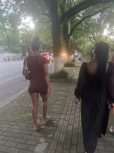 Two women are walking on a paved sidewalk in Liandu District, China. The woman in the foreground is wearing a long-sleeved black dress and has long black braids. The woman in the background is wearing a maroon romper and has her hair in a bun. She is carrying a brown purse. A white car is parked on the side of the road. There are trees and greenery in the background. The lighting suggests it is late afternoon or early evening.