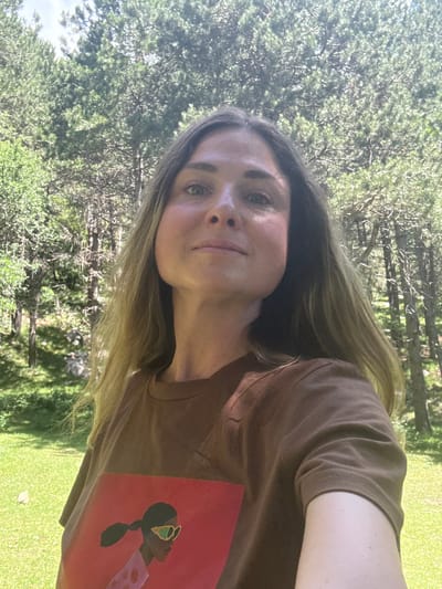 In the village of Žlijebi, Montenegro, a woman with long, light brown hair takes a selfie outdoors. She is wearing a brown t-shirt with a graphic print of a woman wearing sunglasses and a pink polka-dot shirt against a red background. The background consists of lush green grass and tall pine trees, with sunlight filtering through the branches, creating dappled shadows on the ground. The woman's expression is neutral as she looks directly at the camera.