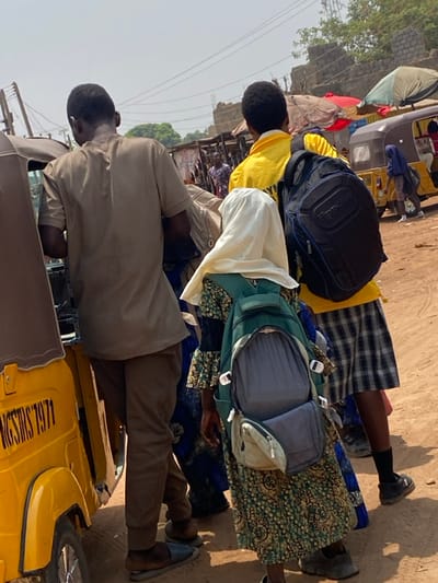 The image depicts an outdoor street or market scene in Katcha, Nigeria, characterized by dry, unpaved ground under a bright, hazy sky. In the immediate foreground, facing away from the viewer, stands a male subject dressed in a light brown short-sleeved top and matching trousers, with dark slippers on his feet. He is positioned next to a yellow three-wheeled auto-rickshaw, identified by the partial registration "NGSMS7971" on its side.

To his right, slightly further back, is a young individual wearing a long, patterned green and blue dress, a white head covering, and a dark green backpack with its front compartment unzipped and open. Adjacent to this individual, also facing away, is another male subject wearing a bright yellow long-sleeved shirt and blue and white plaid shorts. He carries a black backpack with a visible brand logo.

In the mid-ground, the scene continues with more individuals and several market stalls or structures featuring colorful tarpaulin coverings, including red and green. Another yellow three-wheeled auto-rickshaw is visible on the right side of the mid-ground, near a person wearing a blue head covering. The background reveals additional sparse vegetation, utility poles with overhead wires, and unfinished concrete block structures. The overall impression is one of a bustling, dusty urban environment during daylight hours.