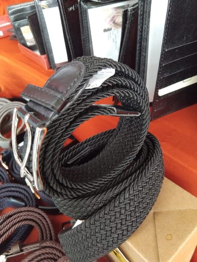This media file shows a close-up, overhead view of several braided fabric belts. The primary subject is a coiled black belt with a silver buckle and a leather keeper. The belt is made of a braided textile that gives it a textured appearance. To the left of the black belt, a black leather wallet is partially visible, open to reveal some cards and a small photograph of a person with blonde hair. In the background, an orange surface is visible, suggesting a display or tablecloth, along with a light brown cardboard box. To the right and lower part of the image, other braided belts in darker colors, possibly dark blue and brown, are also coiled. The lighting suggests an indoor setting, possibly a store or market stall. No specific time of day or weather conditions are indicated. The scene is a still life arrangement of accessories, likely for sale. The location is identified as Vila Nova de Famalicão, Portugal. There is no visible text in the image that is clear enough to read.