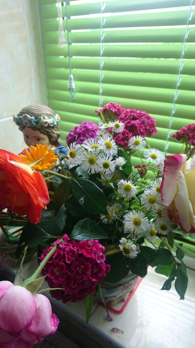 This media file features a vibrant bouquet of flowers, predominantly in shades of pink and white, arranged next to a window with green vertical blinds. The flowers include roses, small daisy-like blooms with yellow centers, and clusters of deep pink carnations. Amongst the flowers, a small figurine of a girl with braided hair adorned with flowers is visible. The scene appears to be indoors, with natural light streaming in from the window. The time of day is likely daytime, given the bright light. There is no visible text in the image. The setting suggests a home environment in Dvirets, Ukraine.