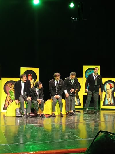 Five men are seated and standing on a brightly lit stage in front of a black backdrop. The men are dressed in formal attire, primarily dark suits and ties. The backdrop features several large, yellow, keyhole-shaped cutouts, each containing a painted illustration. From left to right, the illustrations appear to be a woman in a green dress, a woman with red hair, a man with a yellow tool, and a woman's legs in fishnet stockings and red heels. The stage floor is reflective and green, possibly from stage lighting. The scene appears to be a theatrical performance. The location in Providencia, Chile, is not discernible from the image itself.