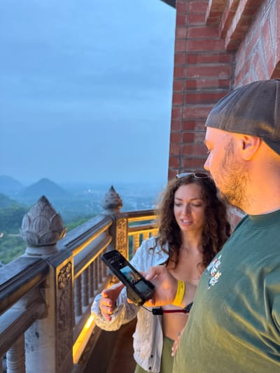 Two people are looking at a smartphone on a balcony in Tây Hoa Lư Ward, Vietnam. The man, wearing a dark baseball cap and a green t-shirt, is on the right side of the frame, his head turned to the left. The woman, with long, curly brown hair, is on the left, wearing a white open shirt over a light-colored top. She holds the smartphone, showing a picture on its screen. Behind them, a stone balustrade with decorative elements overlooks a misty landscape with rolling hills and a city in the distance. The sky is a pale blue, suggesting dusk or dawn. The architecture of the balcony features red brick and ornate stone carvings.