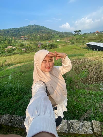 A young woman in a hijab poses for a selfie. She is wearing a beige hijab and a long-sleeved, white, crinkled shirt. She is standing in front of rice terraces on a hillside near Penanggungan, Indonesia. A mountain is visible in the background. The sky is blue.
