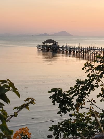 The image depicts a serene sunset scene over the ocean in Labuan Bajo, Indonesia. A long wooden pier extends into the calm water, with a small, thatched-roof structure at its end. Several people are visible on the pier, some standing and appearing to converse or observe the sunset, while others are near the structure. The sky is painted with soft hues of orange and pink, characteristic of a tropical sunset, and distant islands are silhouetted against the horizon. In the foreground, lush green foliage and a cluster of orange flowers add a natural element to the composition, framing the view of the pier and the tranquil sea.