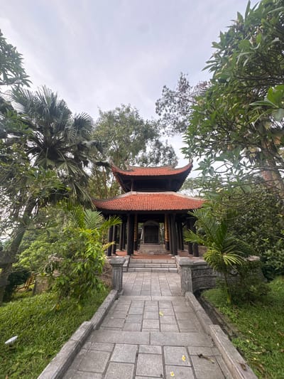 The image shows a traditional Vietnamese temple entrance in Hy Cuong, Vietnam. A paved walkway leads to a temple with a red tiled roof and dark wooden pillars. The temple is surrounded by lush green foliage, including palm trees and other tropical plants. The sky is overcast, suggesting a cloudy day. There are no people visible in the image.