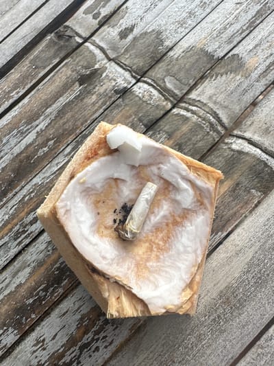 A square-cut coconut husk, acting as an improvised ashtray, is depicted from an overhead angle, resting on a weathered bamboo surface in Tambon Ko Tao, Thailand. The coconut husk has been partially hollowed out, revealing white coconut flesh lining its interior. A partially smoked, hand-rolled cigarette, appearing to be a joint, is vertically inserted into the coconut flesh, with visible black ash and burnt residue surrounding its base. The bamboo surface shows longitudinal patterns and signs of wear and exposure. No individuals are present.