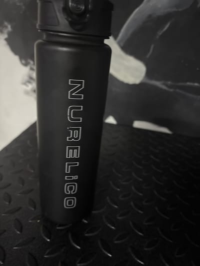 A black reusable water bottle with "NURELICO" inscribed vertically in white capital letters is positioned in the foreground. The bottle is cylindrical with a textured lid. It rests on a black, textured surface, possibly a gym floor or industrial platform, with a raised diamond pattern. In the background, a blurred abstract image, possibly a painting or mural, is visible against a dark wall. The overall lighting is dim, with some highlights on the bottle and the background artwork. The image does not contain any identifiable location information or signs indicating it is in Orsk, Russia.