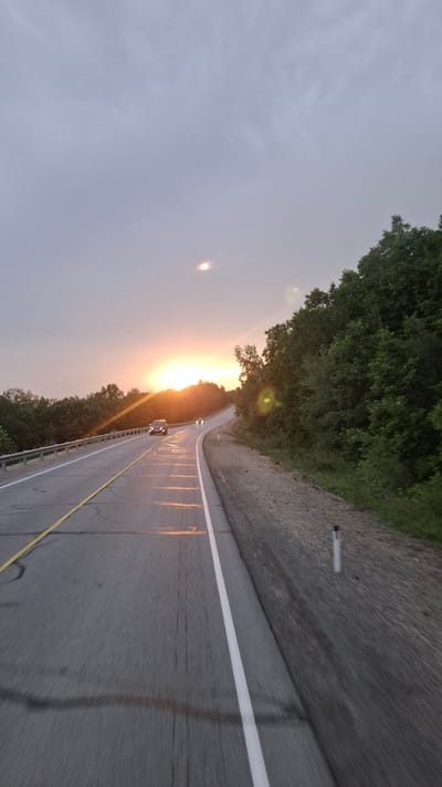 The image depicts a road curving through a wooded area at sunset. The sky is overcast with a soft, diffused light from the setting sun, which appears as a bright, hazy orb near the horizon. The road itself has a yellow line marking the center and a white line on the right edge. There are reflections on the wet asphalt, suggesting recent rain. A silver SUV is visible driving on the road, and two sets of headlights from other vehicles are also present further ahead. Trees line both sides of the road, with dense foliage on the right. The overall impression is one of a serene, natural landscape during twilight, with the road providing a sense of journey or movement. The location is identified as Tarmanchukan, Russia.