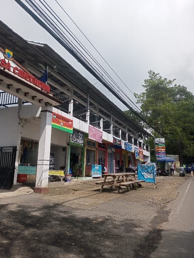 A row of small, two-story commercial buildings line a street. Each building has a shop front with various signs and advertisements, including one for "WARMINDO." A covered walkway runs along the front of the buildings on the second floor. Above the buildings, a tangle of electrical wires is visible against a cloudy sky. A wooden picnic table sits on the sidewalk in front of the shops, and a few people and motorbikes are seen further down the street.