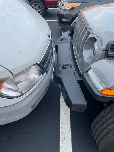 This close-up photograph shows the front ends of two cars parked nose to nose in a parking lot. The upper vehicle, a grey Jeep Wrangler, faces downward, its front bumper practically touching the hood of a white sedan below. The Wrangler features its distinctive seven-slot grille, circular headlights, and an orange side marker light. Part of its front tire is visible in the upper right. The white sedan has a modern headlight design and a chrome-accented grille. The asphalt parking lot is marked with faded white lines, and a partial view of a third vehicle, possibly a red golf cart or small car, is seen on the left. This image was taken in Largo, Florida, United States.