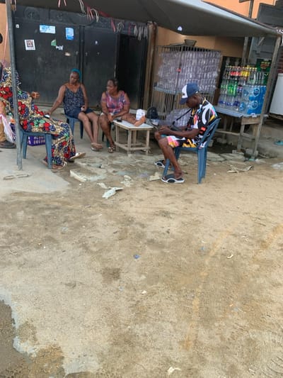 This image depicts an outdoor scene in Ikota, Nigeria, under bright daylight, showing four individuals gathered on an unpaved, sandy ground, likely in front of a small shop or residential area.
In the mid-ground, a man wearing a colorful patterned short-sleeved shirt, shorts, a baseball cap, and sandals is seated on a blue plastic chair. He leans forward, seemingly engaged with objects on a small, light-colored wooden table positioned in front of him. To his left, two women are also seated on blue plastic chairs. The woman closer to the man is dressed in a patterned top and shorts, looking towards him. Next to her, another woman in a dark top and shorts faces slightly away. Partially visible on the far left, a fourth woman wears a long, vibrantly patterned skirt and a dark top, seated on another plastic chair.
The background features a small, informal shop or stall with shelves displaying numerous bottles of water and other beverages. Dark metal gates or shutters are also visible, suggesting a closed or partially enclosed structure. The ground is a mix of sand and dirt, with some small debris scattered. The overall atmosphere appears casual and relaxed, indicative of a typical local gathering or informal commercial setting. No specific text on product labels or other signs is clearly legible.