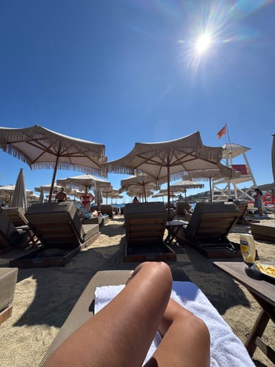 The image captures a sunny beach scene at Agios Ioannis Diakoftis, Greece. The perspective is from a first-person point of view, looking down at the observer's tanned legs resting on a white towel atop a dark brown lounge chair. Adjacent to the observer's right, a small wooden side table holds a yellow Hellmann's mayonnaise bottle and a white plate containing french fries.
The midground features multiple rows of dark brown wooden lounge chairs with light brown cushions, arranged on light-colored sand. Numerous large, hexagonal beach umbrellas with light beige fringed fabric and wooden frames provide shade. Visible individuals include a person partially obscured on a lounge chair directly ahead, a woman in a red bikini top standing between two chairs further back, and several other indistinct figures in the distance.
In the background, a white wooden lifeguard tower with a red base is positioned on the right side of the beach, displaying an orange rectangular flag. Beyond the sandy area, the Aegean Sea is visible, dotted with several boats. A distant landmass featuring white structures is present on the horizon. The sky is clear and bright blue, with a prominent sun flare in the upper right quadrant, indicating intense direct sunlight.