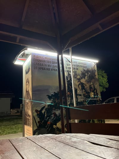 This image captures a night view of a roadside pavilion in Sopot, Bulgaria. The pavilion features a large, illuminated banner with historical imagery and text in Bulgarian. The banner depicts a battle scene with soldiers in historical attire, and portraits. One side of the banner shows a portrait of a woman dressed in traditional clothing, while the other side displays a dynamic scene of armed men in action, with smoke and debris suggesting conflict. Above the imagery, text is visible, with some phrases being "И ДНЕС ИЩ БАЛ", "СПОМНЯ ТОЗ ДЕН", "СЛАВАТА МУ ДИВ", "ОТ УРВА НА УРВА", "ЩУРА ЗАЩА", "ШУ И ПРЕРАЩА", "ТО НА ОИ ЕК", and "О ВЕК НА ВЕК!". The pavilion is supported by metal poles, and a bright LED light strip illuminates the banner. In the foreground, a wooden table is partially visible, suggesting it is part of an outdoor seating area. The background reveals a dimly lit environment with a residential building and grassy area on the left, and a portion of a white vehicle and a green tree on the right. The overall atmosphere is solemn and commemorative.