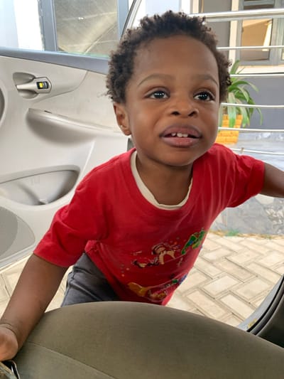A child in a red shirt with cartoon characters is in a car. The child is standing with their arm extended, leaning on the car door. The car door is white and has a speaker grill. The car seat is olive green. The background shows light-colored brick flooring and a plant. The child is looking directly at the camera with a slight smile, and their head is tilted. The child has dark hair and eyes.
