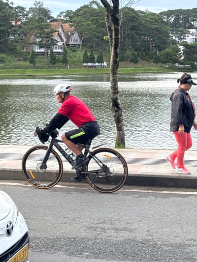 An outdoor, medium shot shows a man cycling on a paved path next to a body of water. The man is wearing a red shirt and black shorts, and a white helmet. He is on a dark-colored road bike. To the right of the cyclist, a woman walks on the paved path. She is wearing a black jacket, red pants, and a black baseball cap. The path is bordered by a body of water to the left of the cyclist and a grassy area with trees and houses to the left of the water. The location is identified as Xuân Hương Lake in Đà Lạt, Vietnam. The foreground shows the front of a white car, partially visible. The sky is overcast.