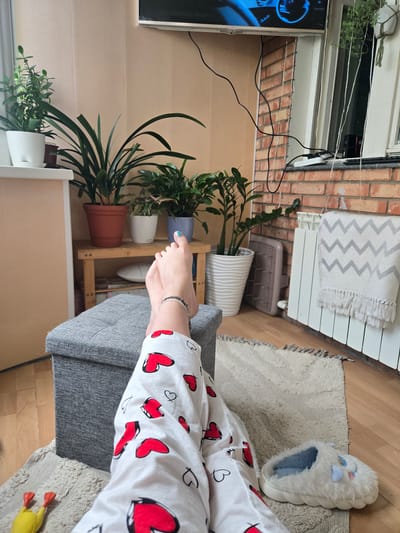 The image shows a pair of feet wearing white pajama pants with red hearts, resting on a grey ottoman. The feet are adorned with a silver anklet. The background features several potted plants, a wooden shelf, a radiator with a patterned blanket draped over it, and a television mounted on the wall. The flooring is wooden, with a textured rug. A fluffy white slipper with blue accents is visible on the rug. This scene is not explicitly located in the city of Kimry, Russia, and no specific details suggest this location.