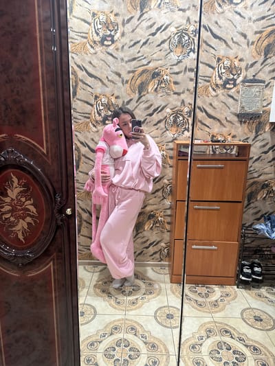 A person is lying on a tiger-print rug, holding a large pink stuffed panther. They are wearing a pink tracksuit and holding a smartphone. The scene appears to be indoors, possibly a bedroom or living room, with a wooden cabinet and tiled floor visible. The lighting suggests daytime. The reflection in what appears to be a mirrored surface shows the person and their surroundings. The backdrop is a tiger print wallpaper. The person's expression is neutral. There is no visible text. The location is Magnitogorsk, Russia.