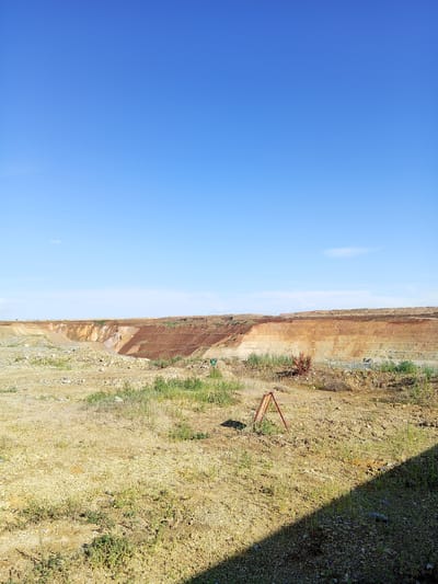 This is a wide-angle, eye-level, outdoor shot of a strip mine under a clear blue sky. The mine itself is a large, multi-layered excavation with steep, reddish-brown walls. The layers are distinct, suggesting different strata of rock or soil have been exposed. The foreground is a desolate, rocky terrain with sparse, dry vegetation, indicative of a harsh or arid climate. A small, red, A-frame marker is visible in the middle ground. The scene appears to be set in the daytime, with bright sunlight casting a distinct shadow across the lower right portion of the image. The overall impression is one of an industrial landscape, possibly in a remote or undeveloped area, given the lack of visible structures or infrastructure beyond the mine itself. The location is identified as Raygorodok, Kazakhstan.