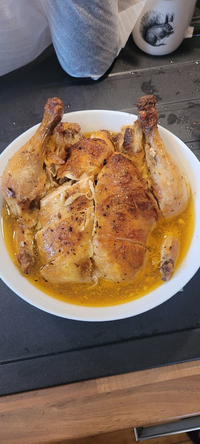The image shows a platter of roasted chicken on a dark countertop. The chicken is golden brown and appears to be seasoned with herbs and spices. It is served in a white bowl with a yellow liquid, likely chicken broth or juices. The background is out of focus but shows what appears to be a squirrel mug and some fabric. The countertop is dark and textured, with some faint water droplets. The overall scene suggests a home kitchen setting. The image does not provide any information to suggest a location in Dietzenbach, Germany.