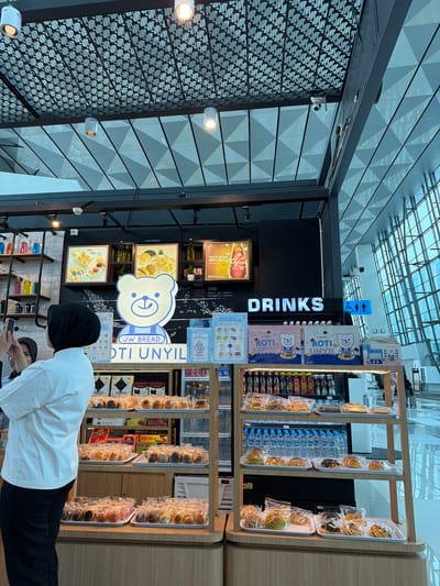 This is a full-shot, eye-level, indoor photograph of a bakery counter at what appears to be an airport terminal. The bakery, branded "JW BREAD" and featuring a large cartoon bear mascot, is well-lit with overhead lights. The counter displays various pastries and bread products, neatly arranged on trays and individually packaged. Shelves behind the counter hold more packaged goods and drinks. In the foreground, a woman wearing a white chef's coat and black headscarf is taking a photo with her phone. The background reveals a modern architectural design with large windows and geometric ceiling patterns. This scene is located in the city of Rawa Burung, Indonesia.