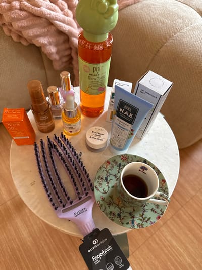 An overhead photograph captures a collection of beauty products, a hairbrush, and a teacup arranged on a round, white marble-patterned side table. The background features a blurry light beige, ribbed fabric, likely upholstery, and a light wooden floor. The scene depicts an indoor domestic environment. No specific geographical markers are present within the image to identify a precise location within Saint-Cyr-l'École, France.

**Objects present on the table, from left to right, top to bottom:**
*   An orange rectangular box for Avene 50+ Intense Protect sunscreen.
*   A brown pump bottle labeled Wella Ultimate Repair Miracle Hair Rescue.
*   A small clear glass bottle containing a light purple liquid, possibly nail polish.
*   A small clear glass dropper bottle with yellow liquid, labeled The Nature of Things Serum Vitamin C.
*   A large orange bottle with a light green Hello Kitty-shaped cap, identified as Pixi Hello Kitty Glow Tonic (8.5 fl oz / 250 ml).
*   A small white lidded jar, labeled Colour & Other Stories Eye Colour Cream.
*   A light blue tube, labeled NAE Hydration & Eclat, certified organic.
*   A white rectangular box standing vertically, with "THE IMPOSSIBLE TAN" printed on its side and "MADE BY ME" on the top flap.
*   A smaller white rectangular box positioned behind the NAE tube, indicating "Huile cuticules et ongles" (Cuticle and nail oil).
*   A lavender-colored Olivia Garden Fingerbrush with black bristles and a black product tag hanging from its handle.
*   A teal-green cup and saucer set with a multi-colored floral and bird pattern. The cup contains a dark brown liquid, consistent with coffee or tea.

No individuals are present in the image. No actions or interactions are depicted; the scene is a static arrangement of items.