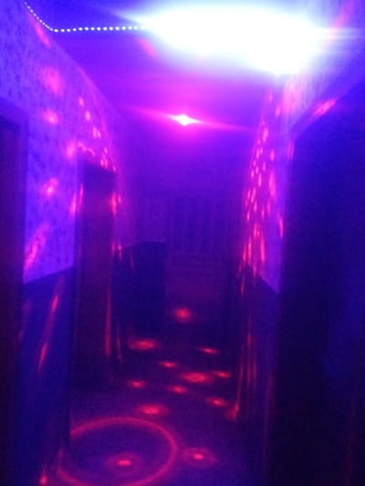 The image depicts an indoor environment, likely a hallway or corridor, characterized by intense purple and blue lighting. The walls are illuminated with scattered pinkish-red spots, suggesting a light effect or projections. A bright white light emanates from the ceiling at the far end of the corridor, casting a strong glow. Red circular patterns and smaller red spots are visible on the floor, possibly from lighting fixtures or projections. The overall scene conveys a vibrant, possibly playful or entertainment-oriented atmosphere. No people or specific actions are discernible. The provided location information, "Sabon Gari Nassarawa, Nigeria," is not visually supported by any elements within the image itself.