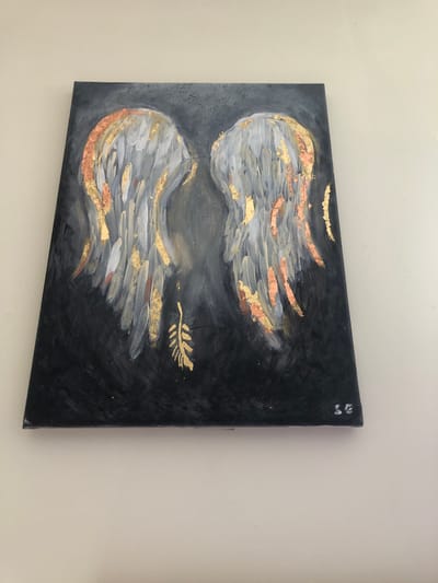 An abstract painting of angel wings is depicted against a dark background. The wings are rendered in shades of grey and white with accents of gold and copper leaf. A single gold feather is painted in the center below the wings. The painting is signed with the initials "LC" in the bottom right corner. The painting is mounted on a white wall. The location of Rother, United Kingdom is not depicted in the image.